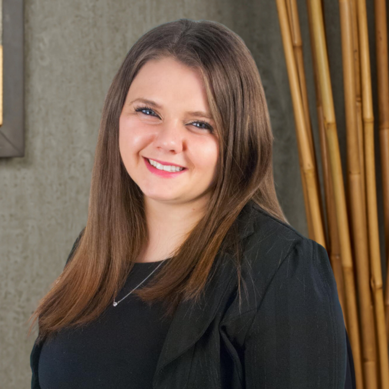 Portrait of a smiling woman with long brown hair, wearing a black blazer against a concrete and bamboo backdrop.