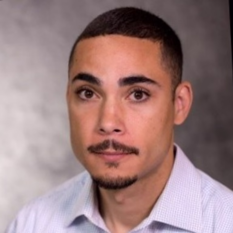 Portrait of a young man with short dark hair and a goatee, wearing a light button-down shirt against a gray backdrop.