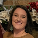 Smiling woman with shoulder-length dark hair in a brown top, holiday greenery and a brick wall behind her.