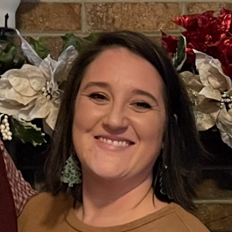 Smiling woman with shoulder-length dark hair in a brown top, holiday greenery and a brick wall behind her.