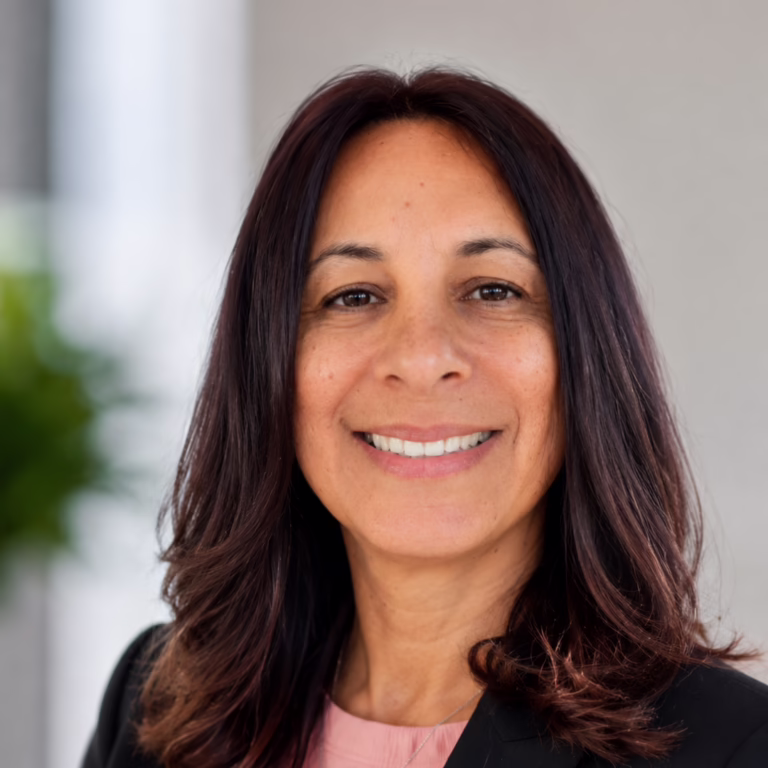 Professional headshot of a smiling woman with shoulder-length dark hair, wearing a pink blouse and black blazer against a neutral background.