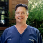 Smiling man in blue medical scrubs outdoors with green foliage in the background.