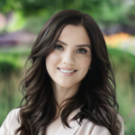 Smiling woman with wavy dark brown hair outdoors, wearing a light blouse, soft greenery in the background.