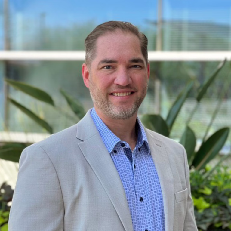 Professional headshot of a smiling man in a light-gray blazer and blue patterned shirt, outdoors with greenery behind.