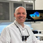 Smiling bald clinician in a white coat wearing dental loupes, in a clinic with a monitor in the background.