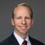 Professional headshot of a smiling man in a dark suit and burgundy tie against a gray gradient background.