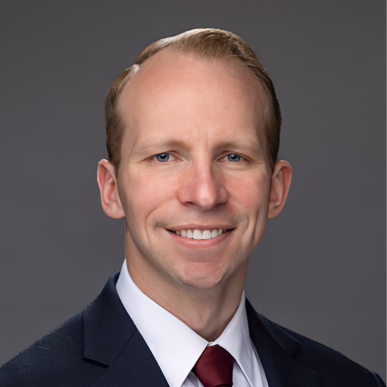 Professional headshot of a smiling man in a dark suit and burgundy tie against a gray gradient background.