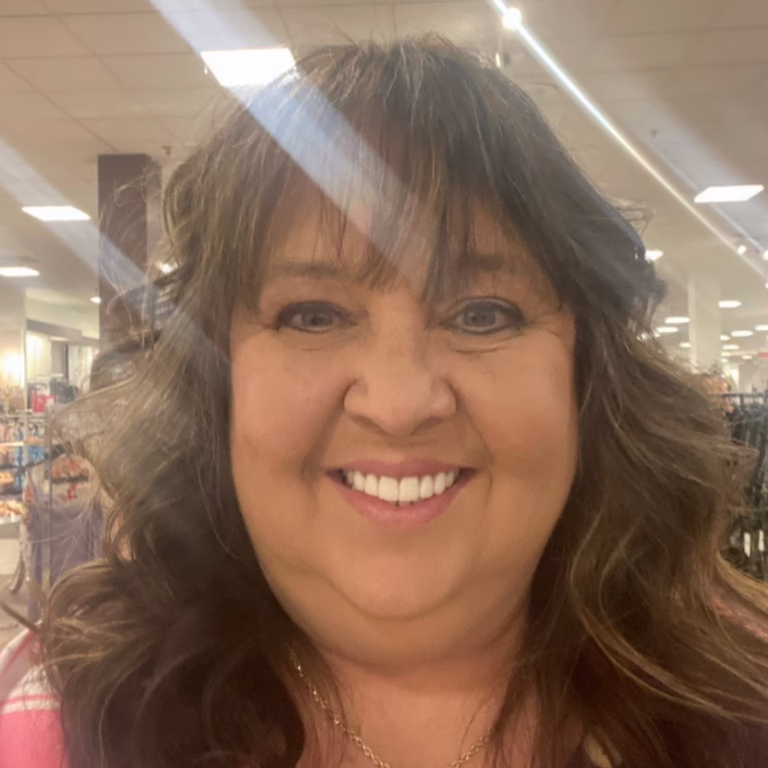 Smiling woman with shoulder-length brown hair in a bright department store, wearing a pink top.