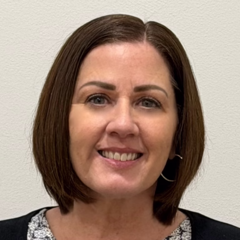 Head-and-shoulders portrait of a woman with shoulder-length brown hair, warm smile, wearing a patterned top and black cardigan against a beige wall center frame.