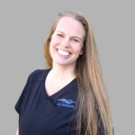 Portrait of a smiling female dentist in navy scrubs with embroidered name on chest, standing against a gray background.