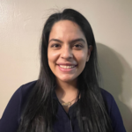 Smiling woman with long black hair wearing a dark blue shirt, standing against a beige wall.