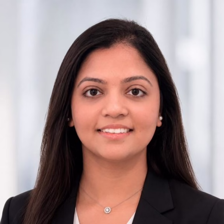 Professional headshot of a woman with long dark hair, wearing a black blazer and pearl earrings, smiling at the camera.