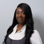Professional headshot of a Black woman with long black hair, wearing a white blouse and gray vest, against a neutral grey background.