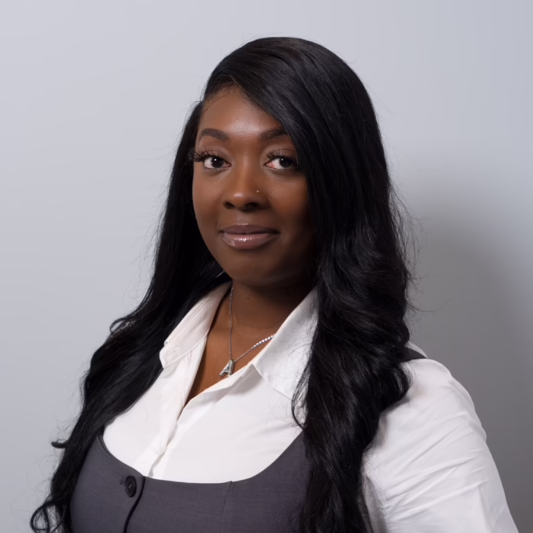 Professional headshot of a Black woman with long black hair, wearing a white blouse and gray vest, against a neutral grey background.