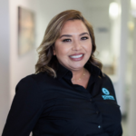 Smiling woman in a black button-down shirt with a teal logo on the chest, standing in a bright hallway (professional portrait).