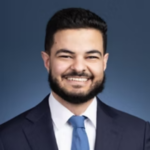 Portrait of a smiling man in a navy suit and light blue tie against a blue gradient background.