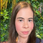 Close-up portrait of a woman with long dark hair and hoop earrings, posed in front of lush green foliage.