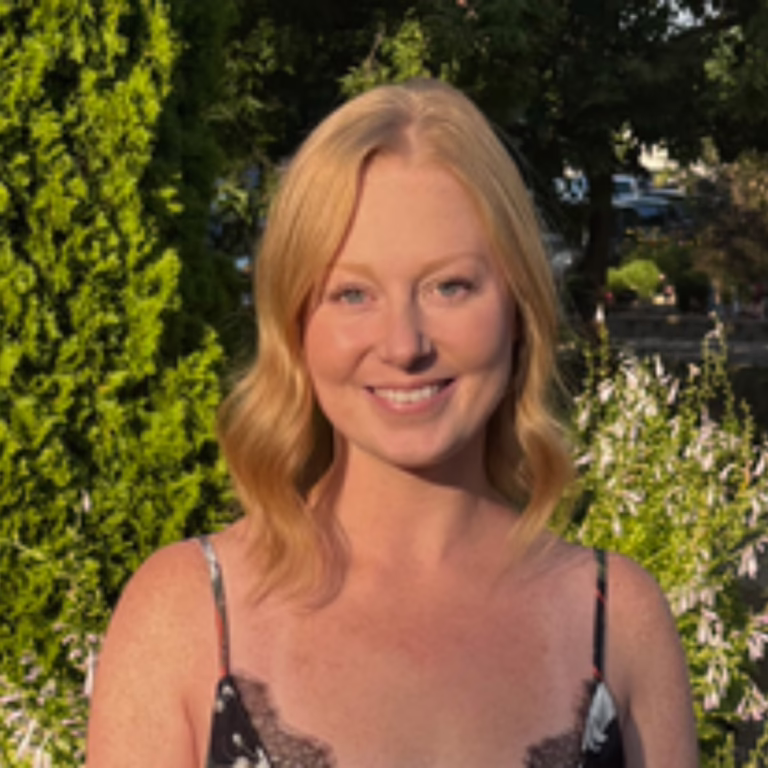 Smiling blonde woman in a black lace-trim dress, outdoors with greenery in the background.