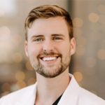 Smiling man with light brown hair and a short beard wearing a white blazer, blurred warm background (professional headshot).