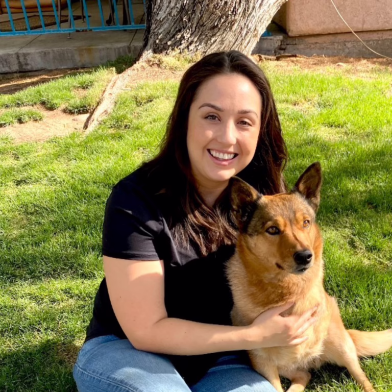 Woman in a black shirt sits on grass with a brown dog, both looking at the camera and smiling outdoors on a sunny day.