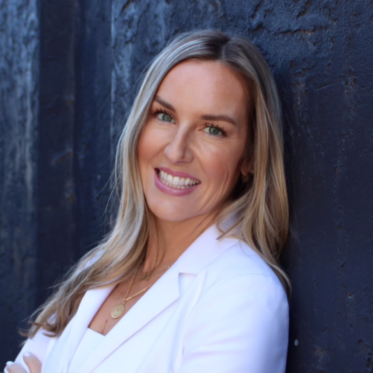 Smiling woman with long blonde hair in a white blazer leaning against a dark blue wall.