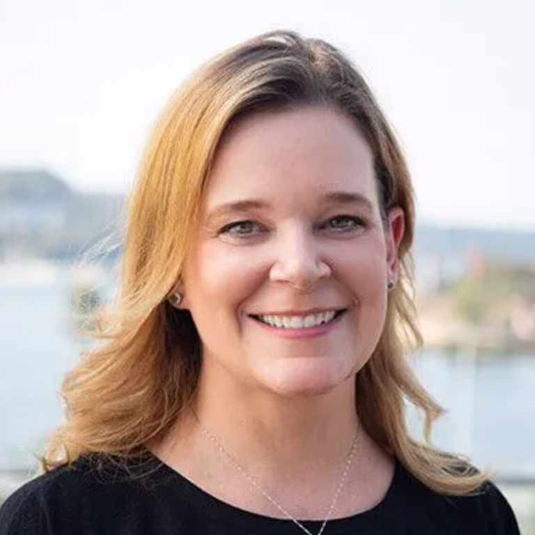 Smiling woman outdoors with a waterfront background, wearing a black top and a delicate necklace.