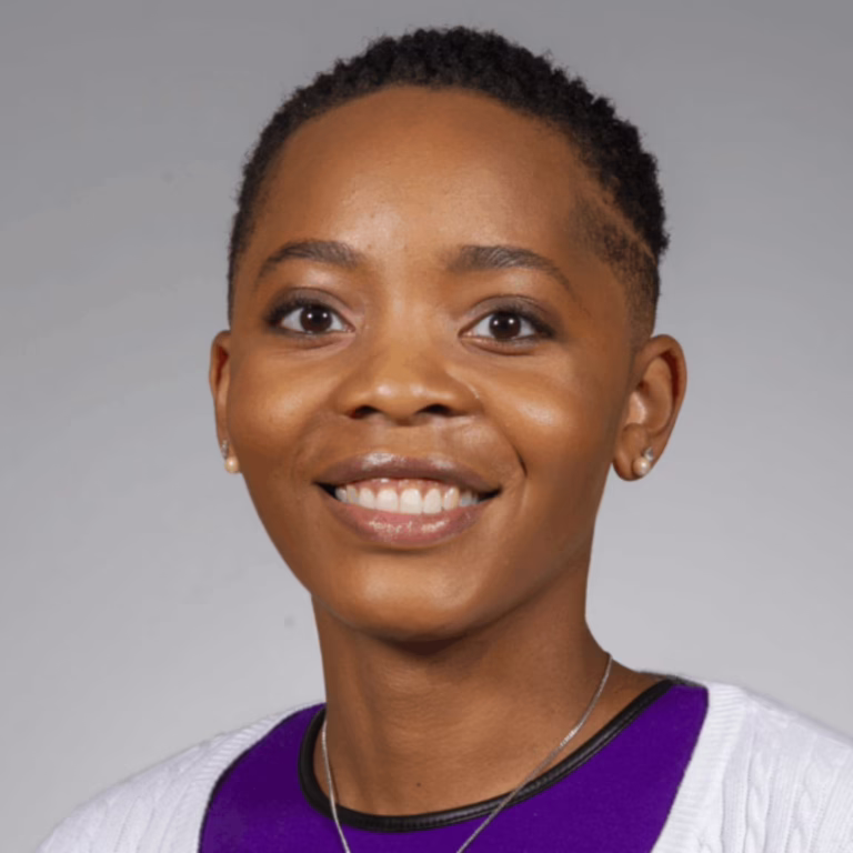Smiling woman with short natural hair wearing a purple top and white cardigan, against a gray background (head-and-shoulders portrait).
