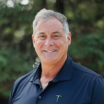 Smiling middle-aged man with gray hair in a navy polo, outdoors with greenery in the background.