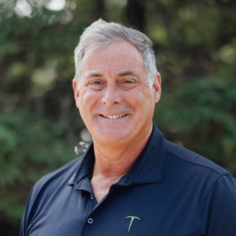 Smiling middle-aged man with gray hair in a navy polo, outdoors with greenery in the background.