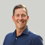 Head-and-shoulders portrait of a smiling man in a navy blue shirt against a neutral gray background.