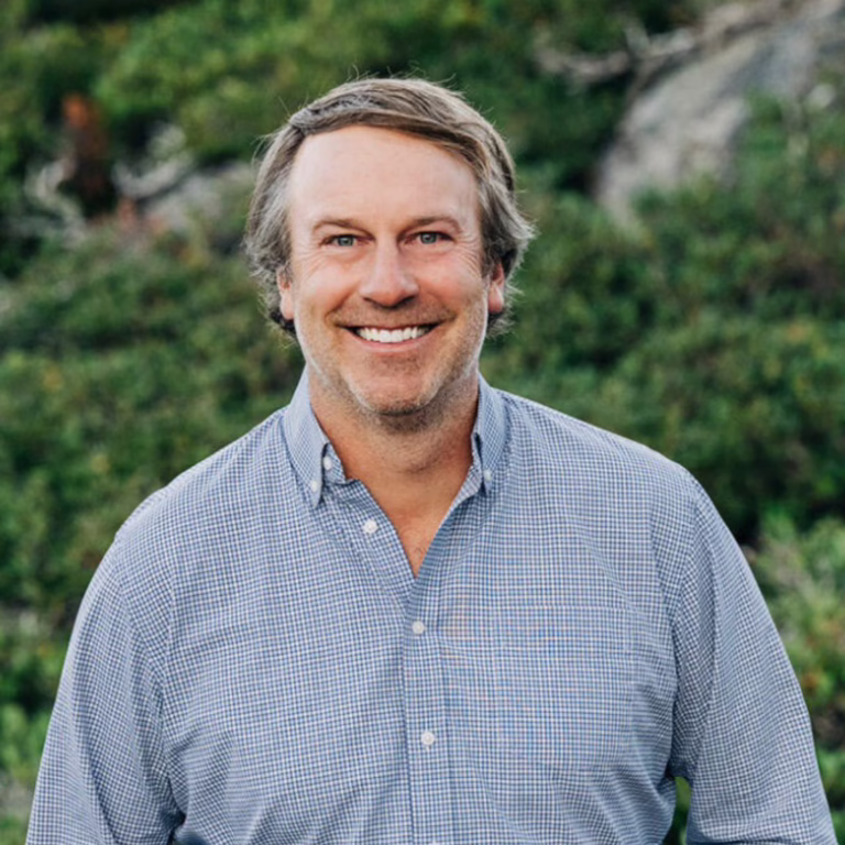 Smiling man in a blue checkered shirt standing outdoors with greenery in the background.