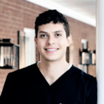 Smiling young healthcare professional in black scrubs posing indoors with a brick wall in the background.