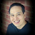 Close-up portrait of a smiling Asian man with short black hair in a dark shirt, posed against a brick wall background.