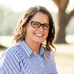 Smiling woman wearing black-framed glasses in a blue-and-white striped shirt outdoors, sunlight in the background.