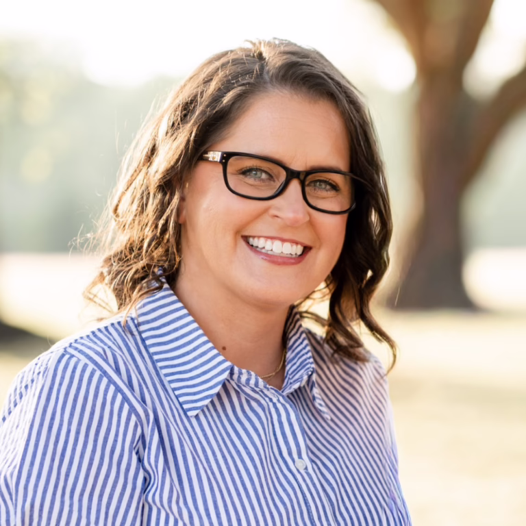 Smiling woman wearing black-framed glasses in a blue-and-white striped shirt outdoors, sunlight in the background.