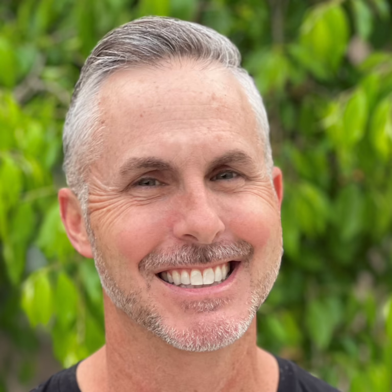 Smiling man with short gray hair and stubble outdoors, green leaves behind.