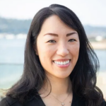 Close-up portrait of a smiling Asian woman with long black hair, wearing a black top and a small pendant, outdoors by the water.