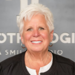 Smiling woman with short white hair and hoop earrings wearing a black jacket and white top, posed against a gray backdrop.