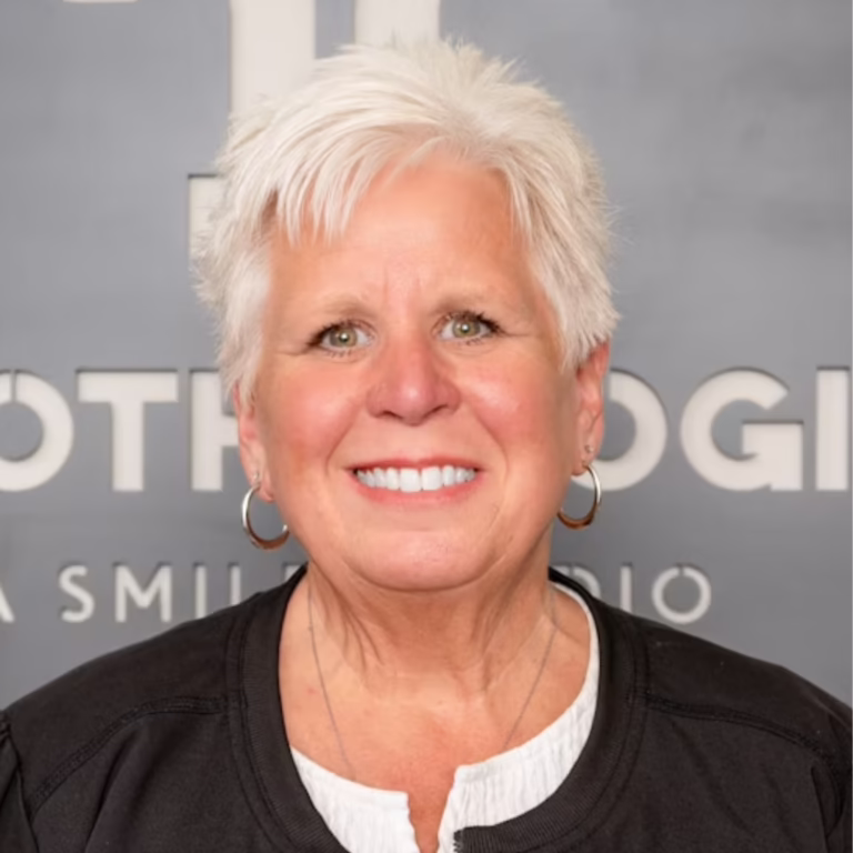 Smiling woman with short white hair and hoop earrings wearing a black jacket and white top, posed against a gray backdrop.