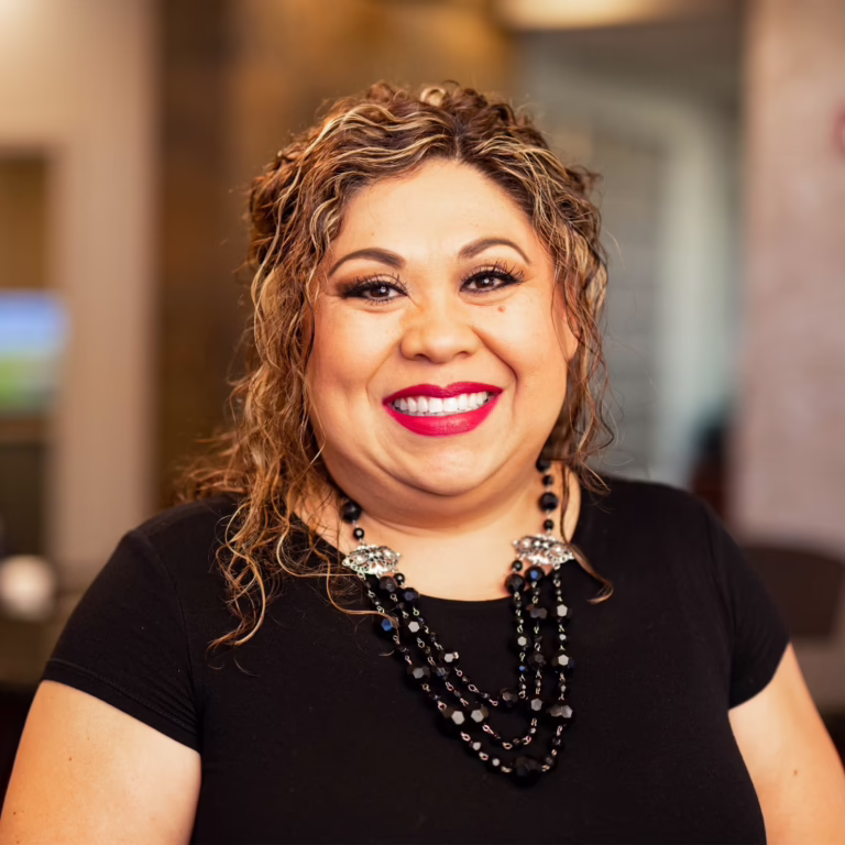 Smiling woman with highlighted curly hair wearing a black top and layered black bead necklace indoors.