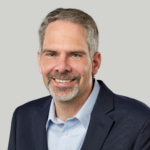 Head-and-shoulders portrait of a smiling man in a navy blazer and light blue dress shirt against a neutral gray background that conveys a professional headshot.