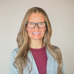 Smiling woman with long wavy blonde hair and black-framed glasses, wearing a light gray blazer over a maroon top, neutral background (professional headshot).