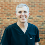 Smiling healthcare professional in navy scrubs against a brick wall backdrop.