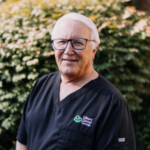 Smiling older man in black scrubs with Gilbert Dental Group logo, outdoors in a garden setting.