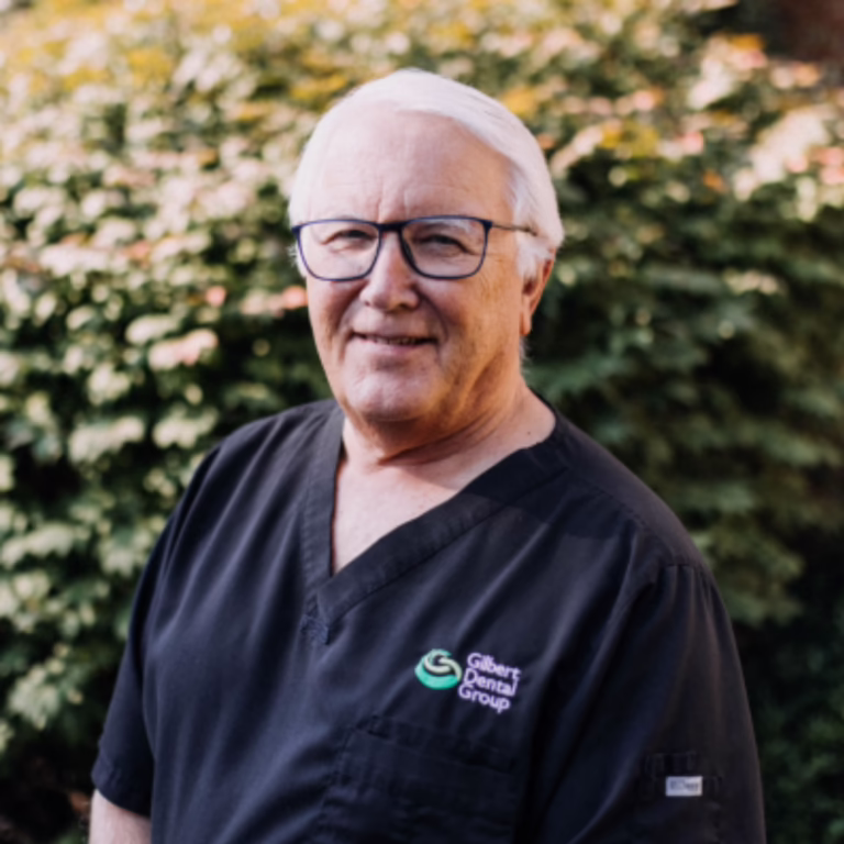 Smiling older man in black scrubs with Gilbert Dental Group logo, outdoors in a garden setting.