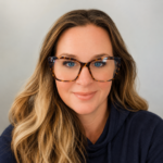 Close-up portrait of a smiling woman with blue eyes and tortoiseshell glasses, wearing a navy hoodie, against a light gray background.