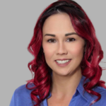 Portrait of a woman with red wavy hair wearing a blue shirt, smiling against a gray background.
