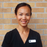 Smiling woman in black scrubs stands against a tan brick wall; name badge on her chest.