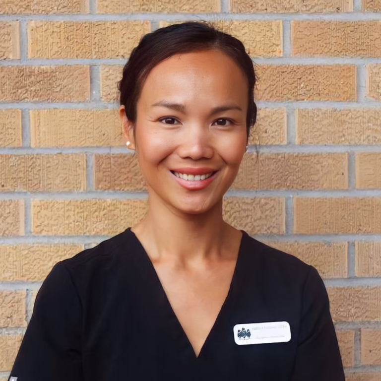 Smiling woman in black scrubs stands against a tan brick wall; name badge on her chest.