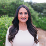 Portrait of a smiling woman with long dark hair, wearing a light blouse and a clover pendant necklace, outdoors with greenery behind her.
